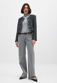 Harmaa tweed-kroppijakki kultaisilla nappuloilla, harmaa poolopaita, vaaleanharmaat leveälahkeiset farkut ja mustat kärjet kengät. Malli seisoo valkoista taustaa vasten.