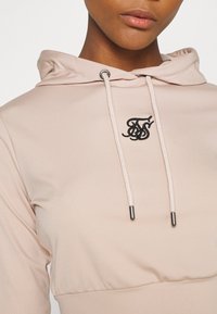 Helle rosa Kapuzenjacke mit Zugbändern mit metallischen Enden und einem gestickten schwarzen Logo auf der Brust. Weicher, strukturierten Stoff.