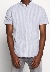 Krátký rukáv košile s knoflíky, s vertikálními modro-bílými pruhy, vyrobená z bavlny. Disponuje sklopeným límečkem a jemnou výšivkou loga.