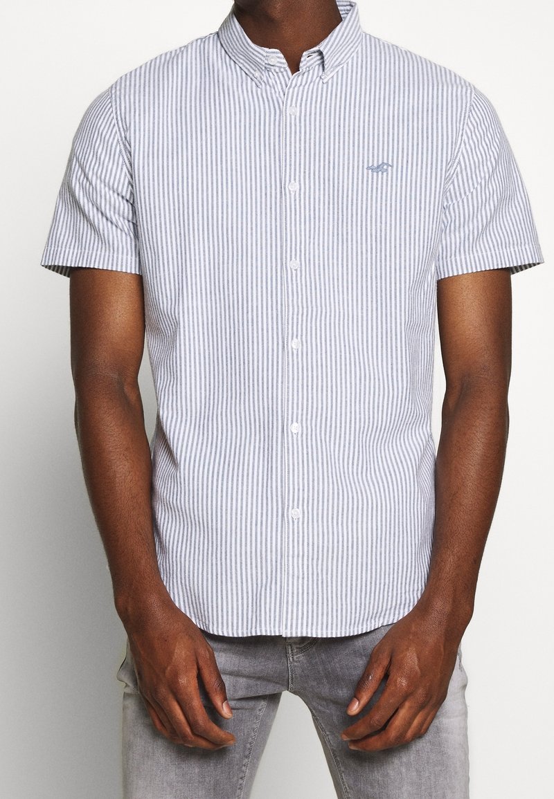 Camisa de manga corta abotonada con rayas verticales azules y blancas, hecha de algodón. Presenta un collar doblado y un sutil bordado del logo.