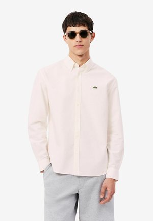 Homme portant une chemise beige clair à boutons avec un petit logo crocodile vert et un pantalon gris, face à l'avant avec une main dans la poche.
