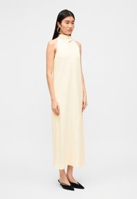 Φόρεμα σατέν με λαιμόκοψη halter σε απαλό κίτρινο χρώμα, μήκος μέχρι το πάτωμα με αέρινη σιλουέτα. Συνδυασμένο με μαύρα mules με μυτερή μύτη για αντίθεση.