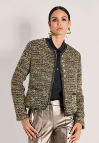 Frau trägt eine strukturierte, mehrfarbige Tweedjacke mit goldenen Knöpfen, kombiniert mit metallisch silbernen Hosen und großen geometrischen Ohrringen.