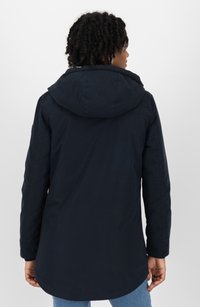 Donna con capelli ricci che indossa una giacca a vento blu scuro con cappuccio, in piedi di spalle contro uno sfondo bianco uniforme.