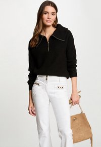 Black zip-up knit sweater with a wide collar, paired with white pants featuring gold accents on front pockets and a tan bag.
