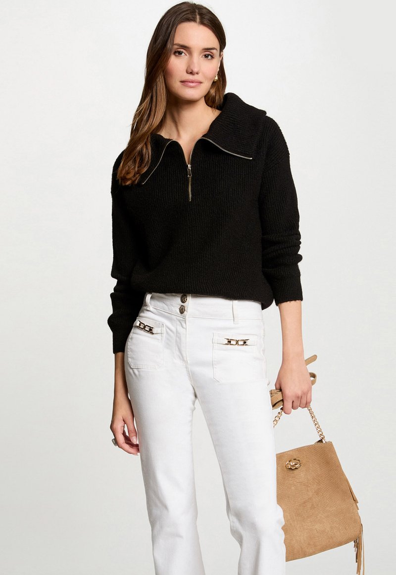 Black zip-up knit sweater with a wide collar, paired with white pants featuring gold accents on front pockets and a tan bag.