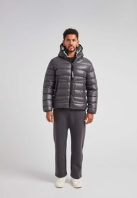 Chaqueta tipo plumón de color gris oscuro brillante con capucha, cierre frontal y dos bolsillos delanteros. Lleva puesta sobre una camiseta negra y pantalones grises, combinada con zapatos blancos.