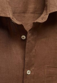 Chemise en lin marron avec un col pointu, une poche poitrine unique et une fermeture avec boutons transparents. Le tissu présente une texture.
