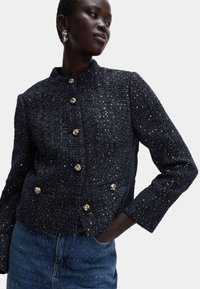 Giacca di tweed navy con finitura testurizzata, caratterizzata da bottoni dorati e colletto rialzato. Abbinata a una gonna di jeans.