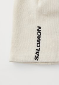Berretto in maglia crema con texture a coste, caratterizzato da un logo ricamato nero ben visibile su un lato.