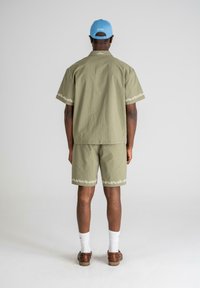 Kortermet olivenfarget skjorte og matchende shorts med blomsterbroderi på mansjettene og kanten, brukt med brune sko og hvite sokker.