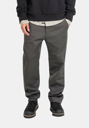 Mann trägt dunkelgraue Hose, schwarze Stiefel, schwarzen Pullover und hellbeiges Hemd, steht mit den Händen in den Taschen vor weißem Hintergrund.