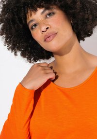 Langdurige mouw oranje top met een ronde hals, met een zachte textuur en een relaxte pasvorm. Close-up van een hand nabij de kraag.