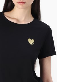 Crna majica s kratkim rukavima izrađena od mekog materijala, s dizajnom s uvijenim rukavima. Zlato-glitter logo srca s natpisom "ONLY" nalazi se na lijevoj strani prsnog koša.