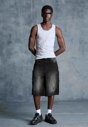 Young man wearing sunglasses, white tank top, long black denim shorts, white socks, and black sneakers standing against a gray textured wall.