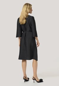 Robe noire à manches trois-quarts, en tissu texturé, taille cintrée et longueur au genou. Portée avec des escarpins noirs à bride arrière ornés de fleurs.