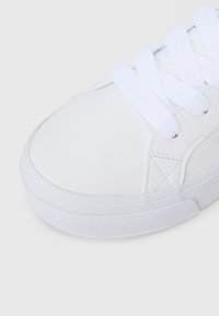 Λευκό δερμάτινο αθλητικό παπούτσι με επίπεδα λευκά κορδόνια, στρογγυλή μύτη, υφασμένη ελαστική σόλα και ελάχιστες ραφές στο πάνω μέρος.