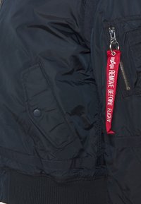 Navy plavi bomber jakna s glatkom teksturom, elastičnim manžetama, džepom na zip i crvenom oznakom "UKLONITI PRIJE LETA".
