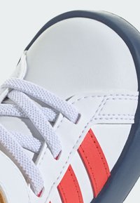 Sneaker bianca in pelle con strisce rosse, lacci bianchi testurizzati e suola in gomma blu scuro. Presenta dettagli cuciti e una punta arrotondata.