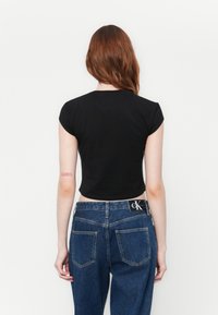 Persoon met lang rood haar, gekleed in een zwart cropped t-shirt en blauwe spijkerbroek, met de rug naar de camera, tegen een eenvoudige witte achtergrond.