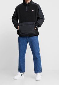 Sudadera negra de forro polar con cuello abotonado, bolsillo frontal y paneles de tela en contraste. Combinada con vaqueros azules de corte amplio y zapatillas blancas.