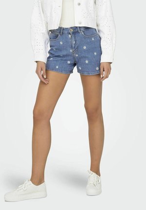 Femme portant un short en denim bleu avec de petits imprimés de marguerites blanches, des baskets blanches et une veste blanche boutonnée avec des détails œillets.