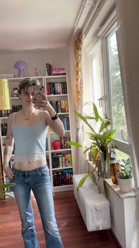 Lyseblå crop-top med blonderkant, kombinert med utslåtte blå jeans. Rommet har bokhyller, planter, og naturlig lys fra et vindu.