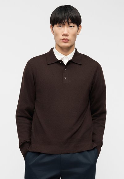 Καφέ πουλόβερ με γιακά και ρεβέρ με δύο κουμπιά. Διαθέτει μακριά μανίκια και πλεκτό ύφασμα με υφή. Συνδυάζεται με μπλε παντελόνι.