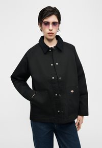 Άτομο με μαύρο σακάκι με κουμπιά και μεγάλες μπροστινές τσέπες και μικρό λογότυπο, μπλε τζιν και γυαλιά ηλίου με μωβ απόχρωση μπροστά σε απλό φόντο.