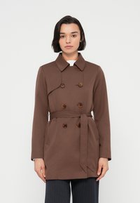 Καφέ παλτό τύπου τραντς με διπλό πέτο, μεγάλα κουμπιά, φαρδύ γιακά, μακριά μανίκια και ζώνη στη μέση.