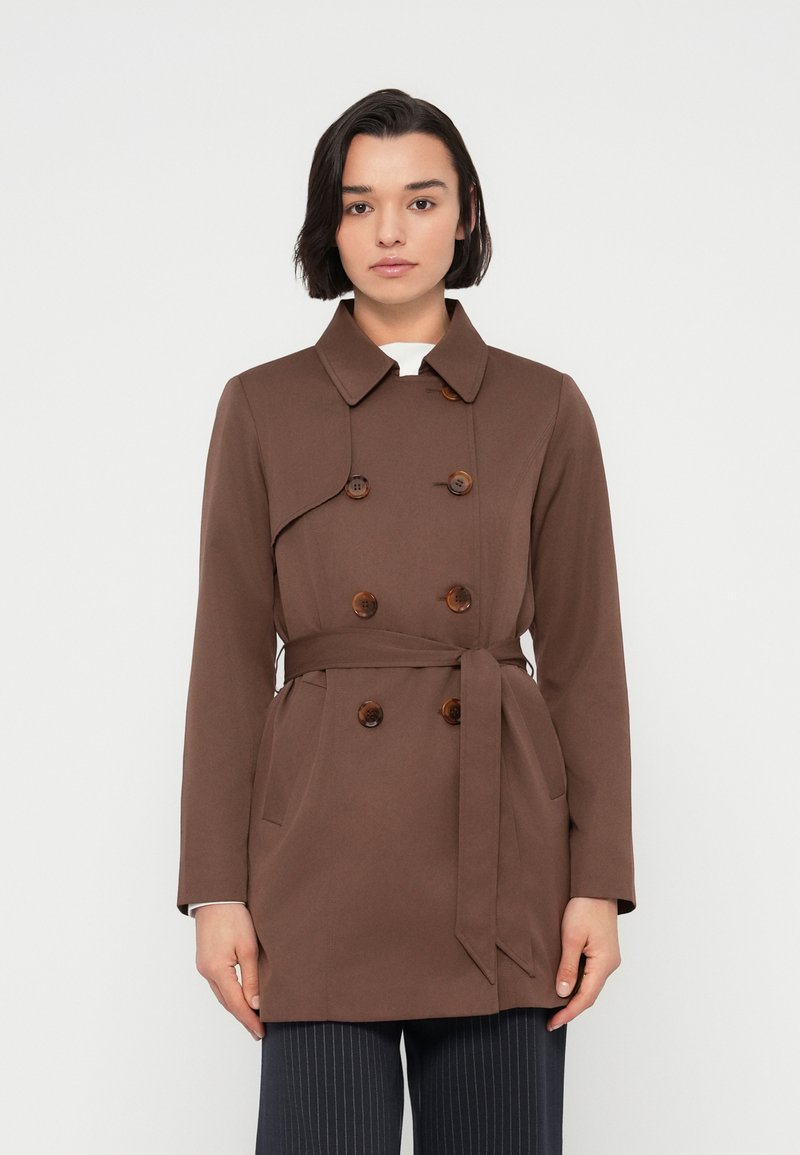 Καφέ παλτό τύπου τραντς με διπλό πέτο, μεγάλα κουμπιά, φαρδύ γιακά, μακριά μανίκια και ζώνη στη μέση.