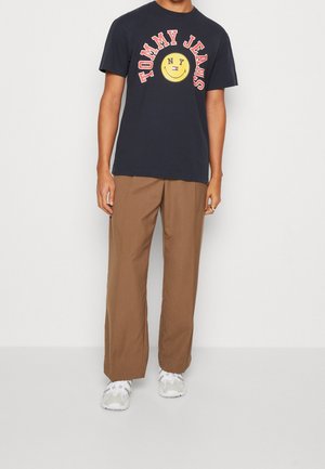 Laivastonsininen puuvillainen t-paita, jossa on punaisella teksti "TOMMY JEANS NY" ja hymyilevä emoiji, yhdistettynä ruskeisiin leveälahkeisiin housuihin ja valkoisiin lenkkareihin.