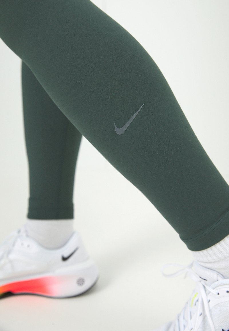 Leggings sportivi verdi realizzati in materiale elastico, con un logo Nike nero sulla coscia, abbinati a sneakers bianche con suole colorate.
