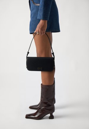 Black leather handbag with a short strap, featuring gold-tone hardware, rectangular shape, and smooth texture, held beside knee-high brown boots.