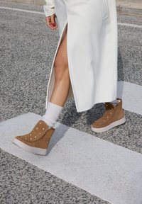 Botas de cuña de ante marrón con tachuelas doradas, suela blanca y costuras laterales, combinadas con calcetines blancos y una prenda larga blanca con una abertura.