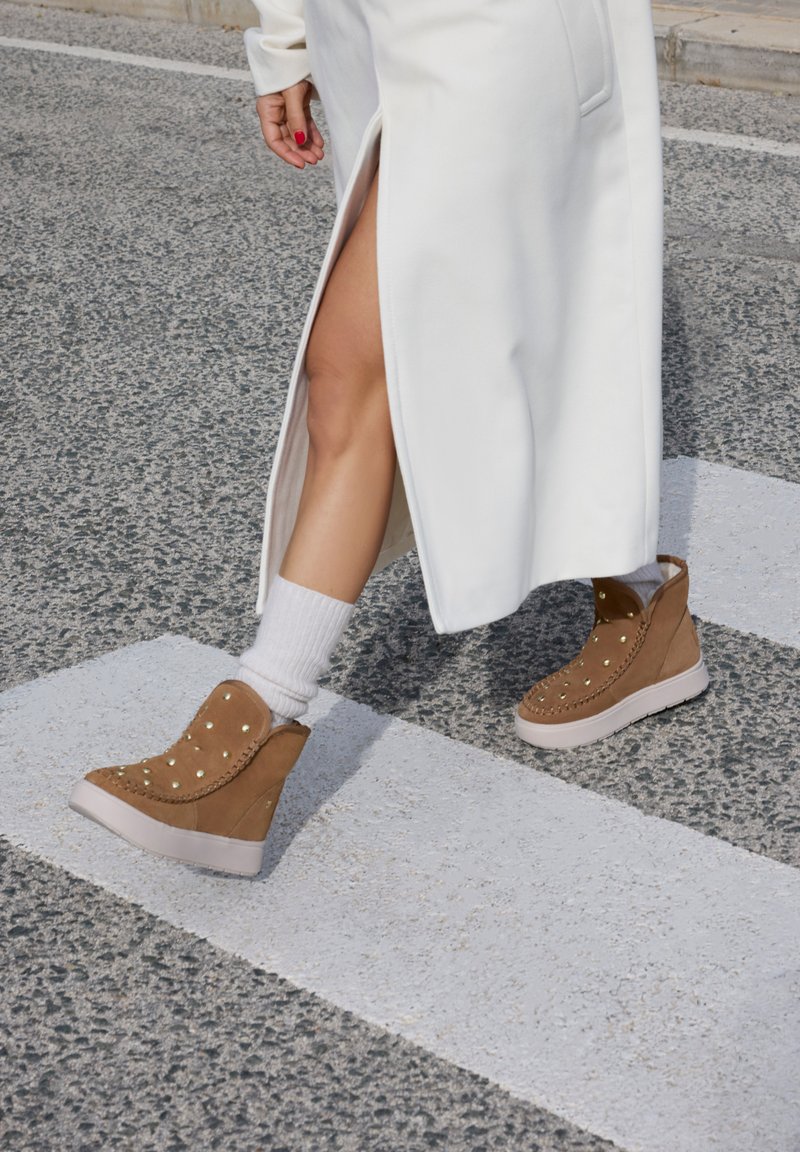 Botas de cuña de ante marrón con tachuelas doradas, suela blanca y costuras laterales, combinadas con calcetines blancos y una prenda larga blanca con una abertura.