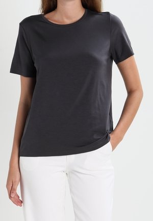 Frau trägt ein schlichtes dunkelgraues T-Shirt mit kurzen Ärmeln und weiße Hose, eine Hand in der Tasche, vor neutralem Hintergrund.