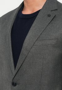 Man in een donkergrijs blazer met zwarte knopen en een donkermarineblauwe ronde hals t-shirt zichtbaar eronder.