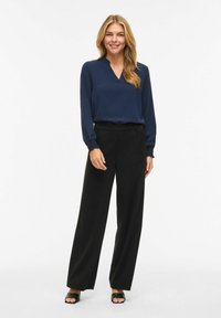 Femme souriante debout, portant un chemisier bleu marine à manches longues, un pantalon large noir et des talons ouverts noirs sur fond blanc.