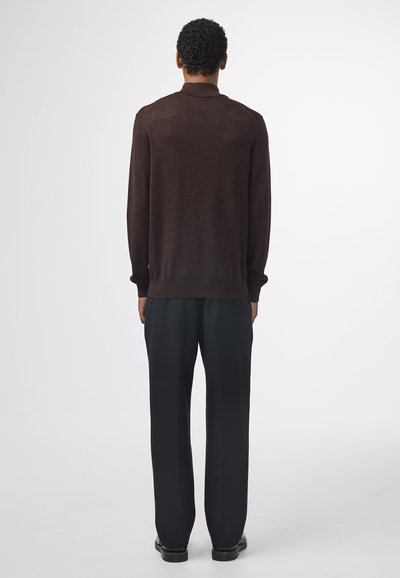 Brauner Strickpulli mit Kragen, langen Ärmeln und gerippten Manschetten, kombiniert mit schwarzen maßgeschneiderten Hosen und schwarzen Schuhen, von hinten betrachtet.