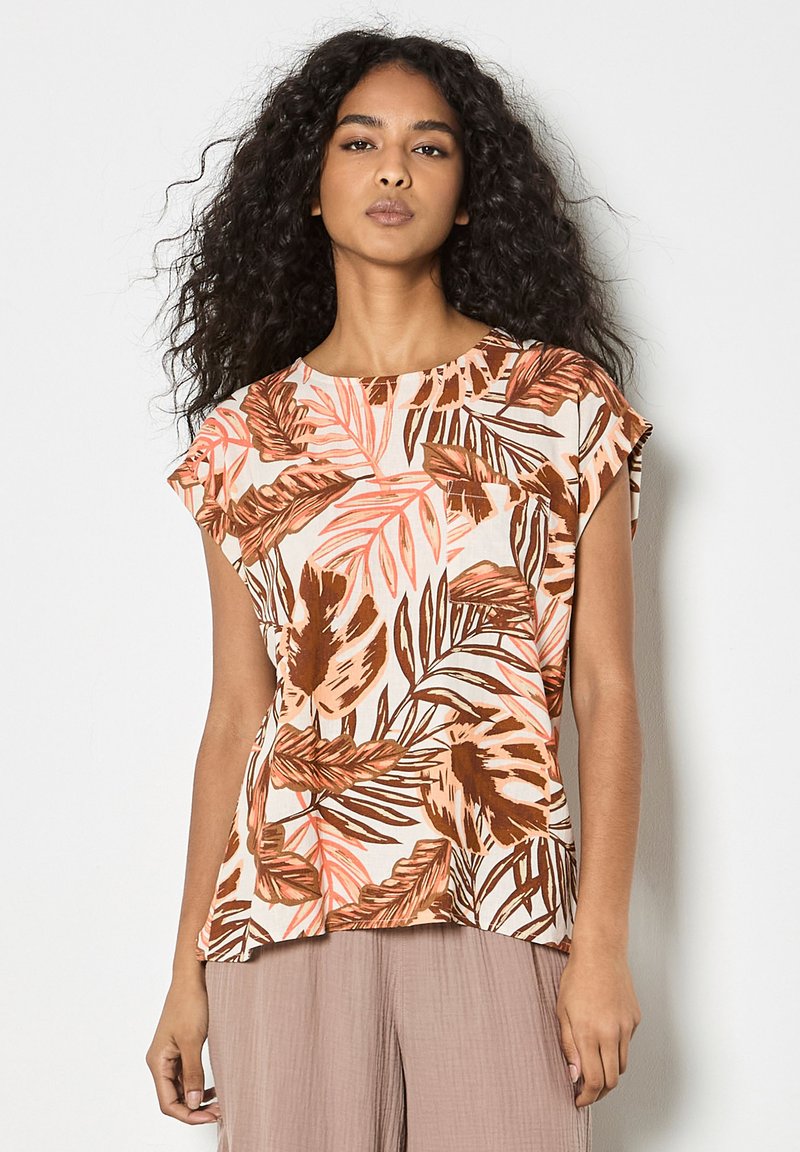 Femme aux longs cheveux bouclés portant un haut sans manches à imprimé tropical en tons marron et pêche avec un pantalon mauve clair.