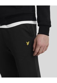 Sorte bomuldsjoggers med sidelommer, der har et lille broderet gult ørneresumé på venstre lår. Tekstureret stofdetalje synlig.