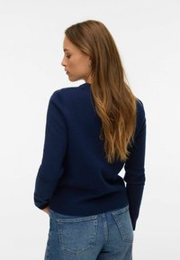 Tumši zila džempera ar apaļu kakla izgriezumu un garām piedurknēm, kopā ar gaiši zilām džinsa biksēm. Gluda tekstūra, pieguļošs stils un ribots apakšmala.