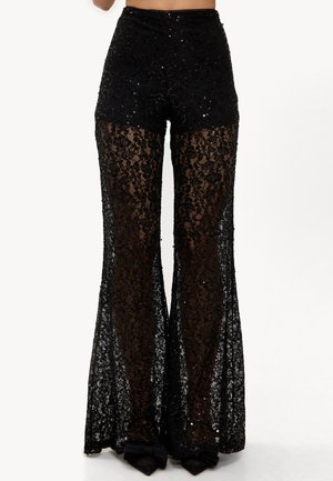 Pantalon noir en dentelle taille haute avec jambes évasées et accents de sequins, porté sur la peau, accompagné de chaussures noires pointues avec des nœuds.