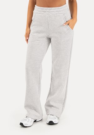 Kvinde iført lysegrå vidde sweatpants med elastisk talje og hvide sportssko, stående mod en ensfarvet baggrund.