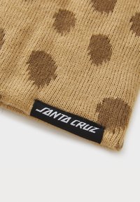 Bruine polka-dot beanie van gebreide stof, met een zwart label waarop "Santa Cruz" in witte letters aan de rand staat.