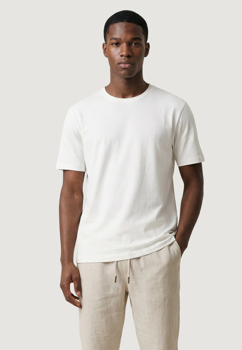 Giovane uomo che indossa una t-shirt bianca a maniche corte semplice e pantaloni beige chiaro con coulisse, in piedi davanti a uno sfondo neutro.