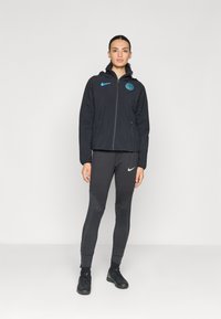 Black zip-up jacket with hood, featuring blue logo and pockets. Black leggings with textured accents and fitted silhouette. Black athletic shoes.