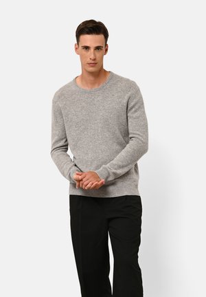 Maglione grigio chiaro, strutturato, con scollo rotondo e polsini a coste, abbinato a pantaloni neri. Modello in piedi con le mani incrociate.