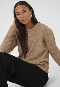 Bruine gestructureerde trui met een ronde hals, gedragen met losse zwarte broek. Model heeft lang haar en zit met een hand om haar nek.